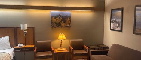 A hotel room with a bed, a desk with a lamp, two chairs, a sofa, and a coffee table. There are framed pictures on the wall.
