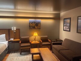 Habitación de hotel con cama, escritorio con lámpara, dos sillas, sofá y una mesa de centro. Hay cuadros en la pared.
