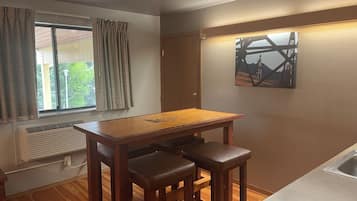 A small kitchen with a wooden table, two stools, a window with curtains, a radiator, and a painting on the wall.