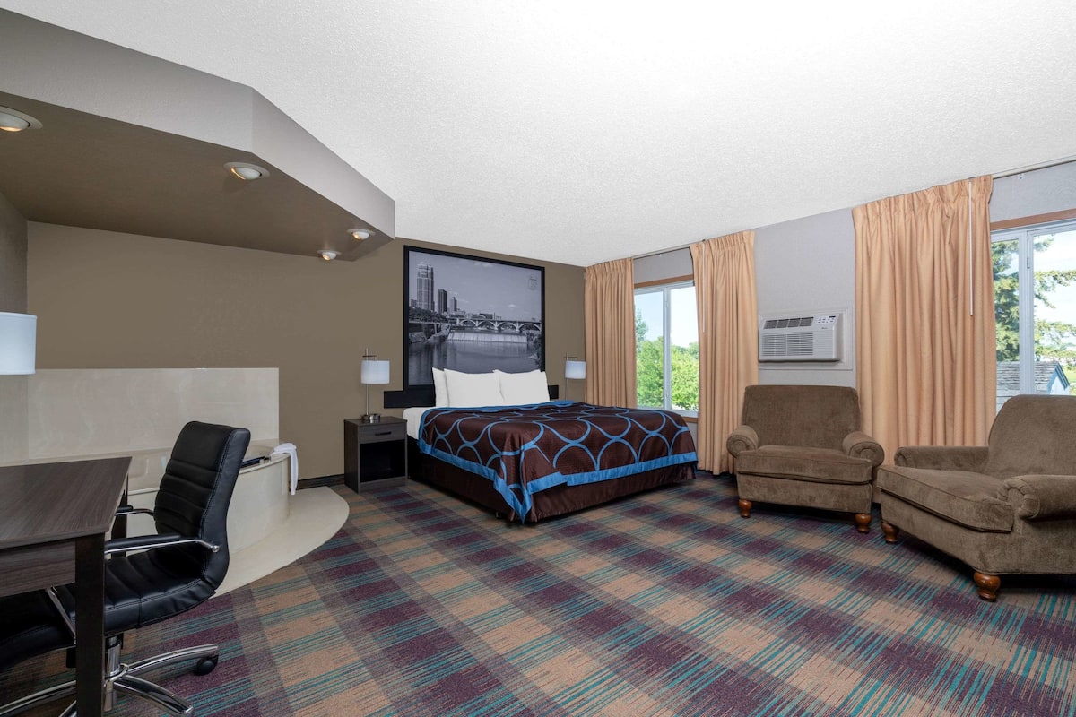 A hotel room with a bed, two armchairs, a desk with a chair, a nightstand, and a large window with curtains.