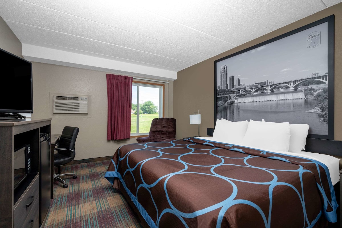 A hotel room with a large bed, a desk with a television, a chair, a window with a view of a bridge, and an air conditioning unit.