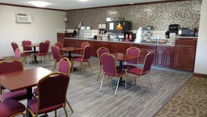 A hotel dining area with red chairs and tables, a kitchen with a microwave and coffee machine, and a patterned wall.
