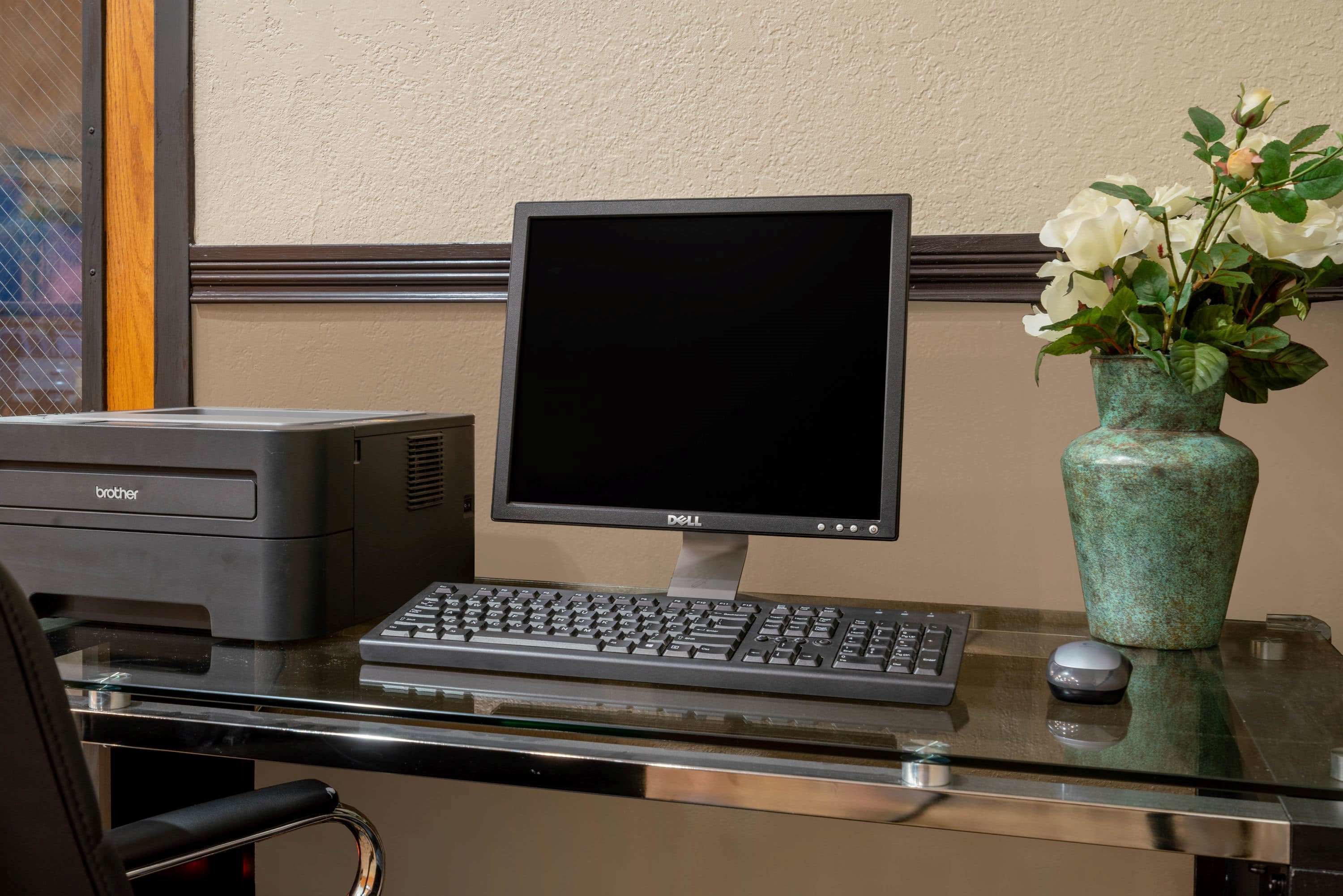 Un escritorio con una impresora Brother, un monitor Dell, un teclado, un ratón y una jarra con flores.