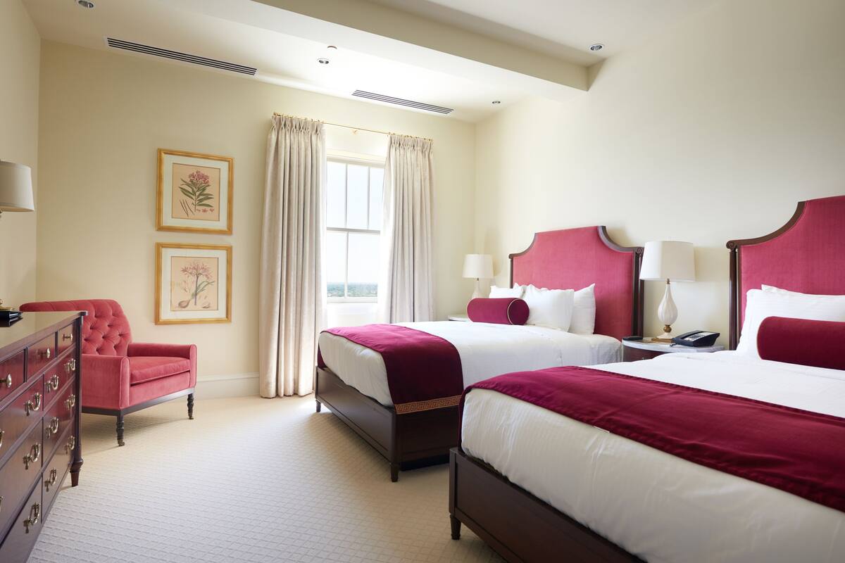 A hotel room with two beds, a red armchair, a dresser, and framed botanical prints on the wall.