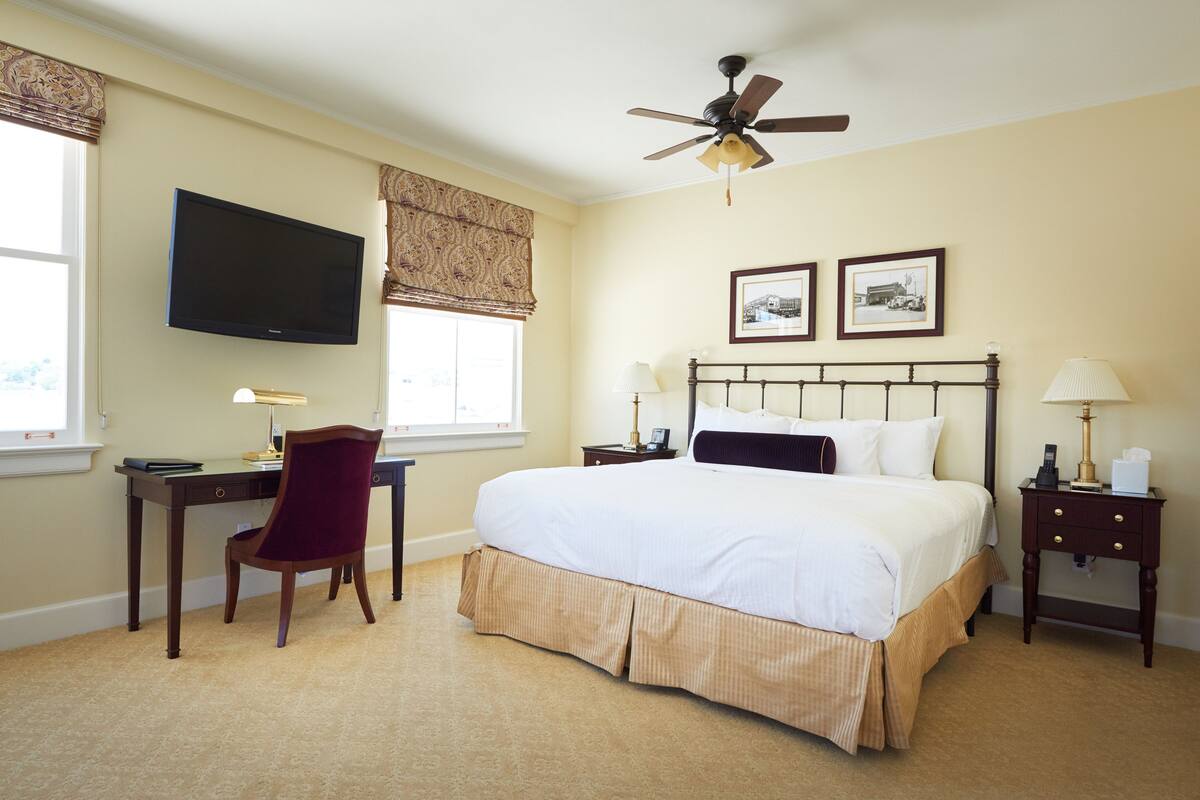 A hotel room with a bed, desk, chair, television, ceiling fan, and two framed pictures on the wall.
