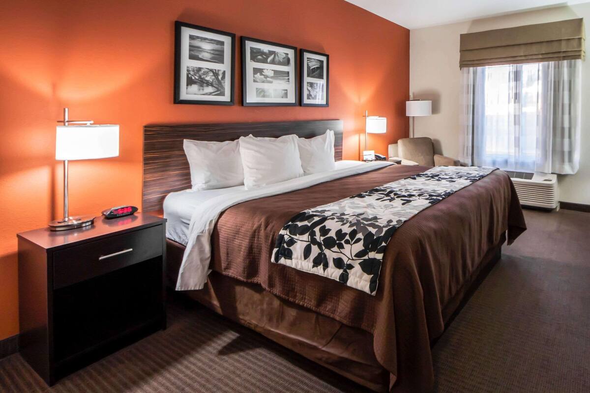 A hotel room with a large bed, a nightstand, a lamp, a chair, and a window with curtains.