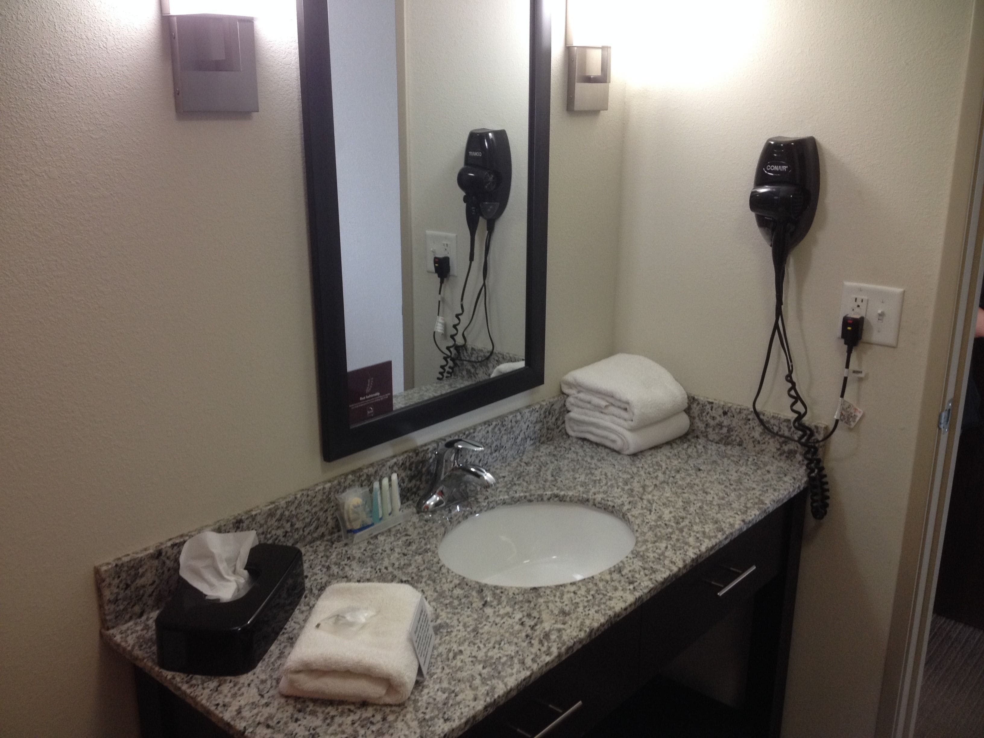 A bathroom with a granite countertop, a white sink, a mirror, a hairdryer, and a towel.