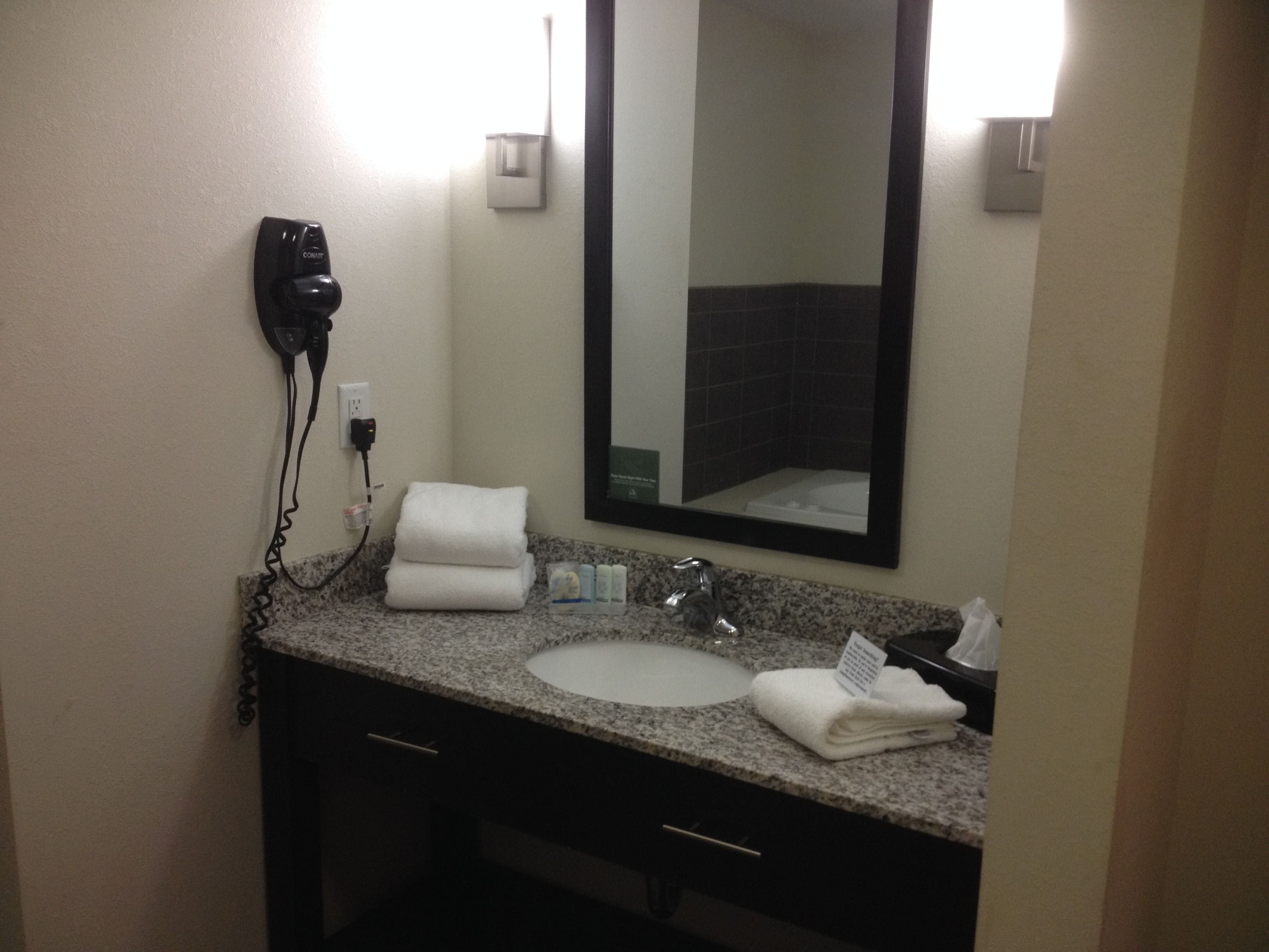 Un baño de hotel con encimera de granito, un lavamanos, un espejo, un secador de pelo y un perchero para toallas.