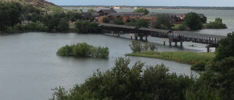 A lakeside view with a wooden bridge connecting two areas, surrounded by greenery and buildings.