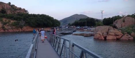 A wooden bridge over a river with people walking, cars parked nearby, and a mountain in the background.