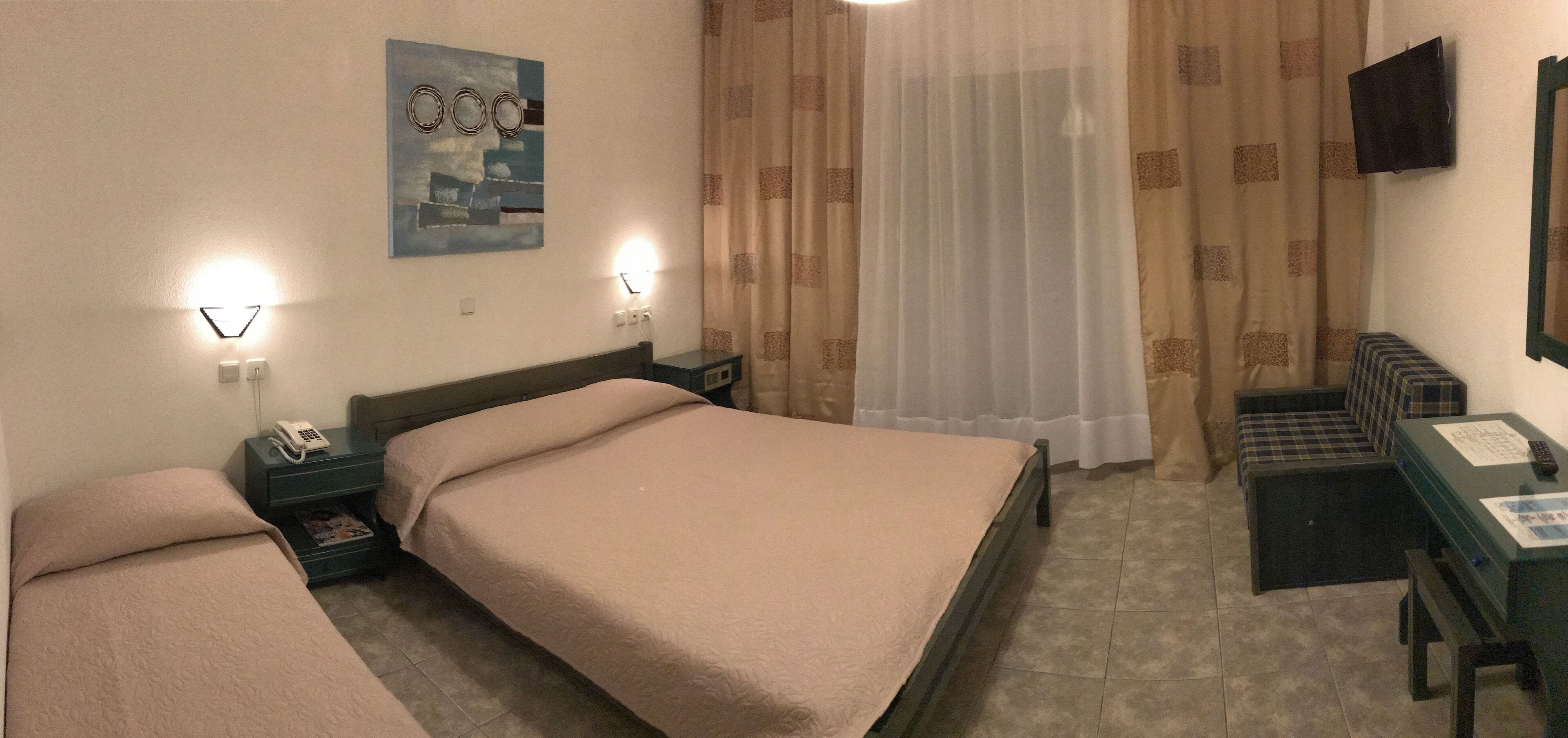 A hotel room with two single beds, a desk with a chair, a telephone, a television, and a window with curtains.