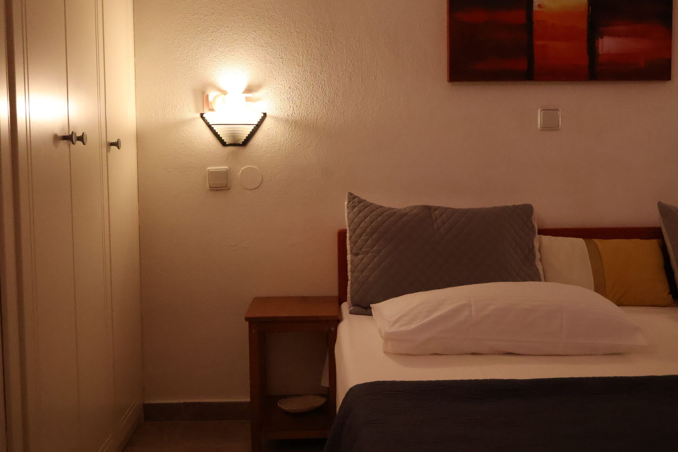 A hotel room with a bed, a nightstand, a wall-mounted light, and a door.
