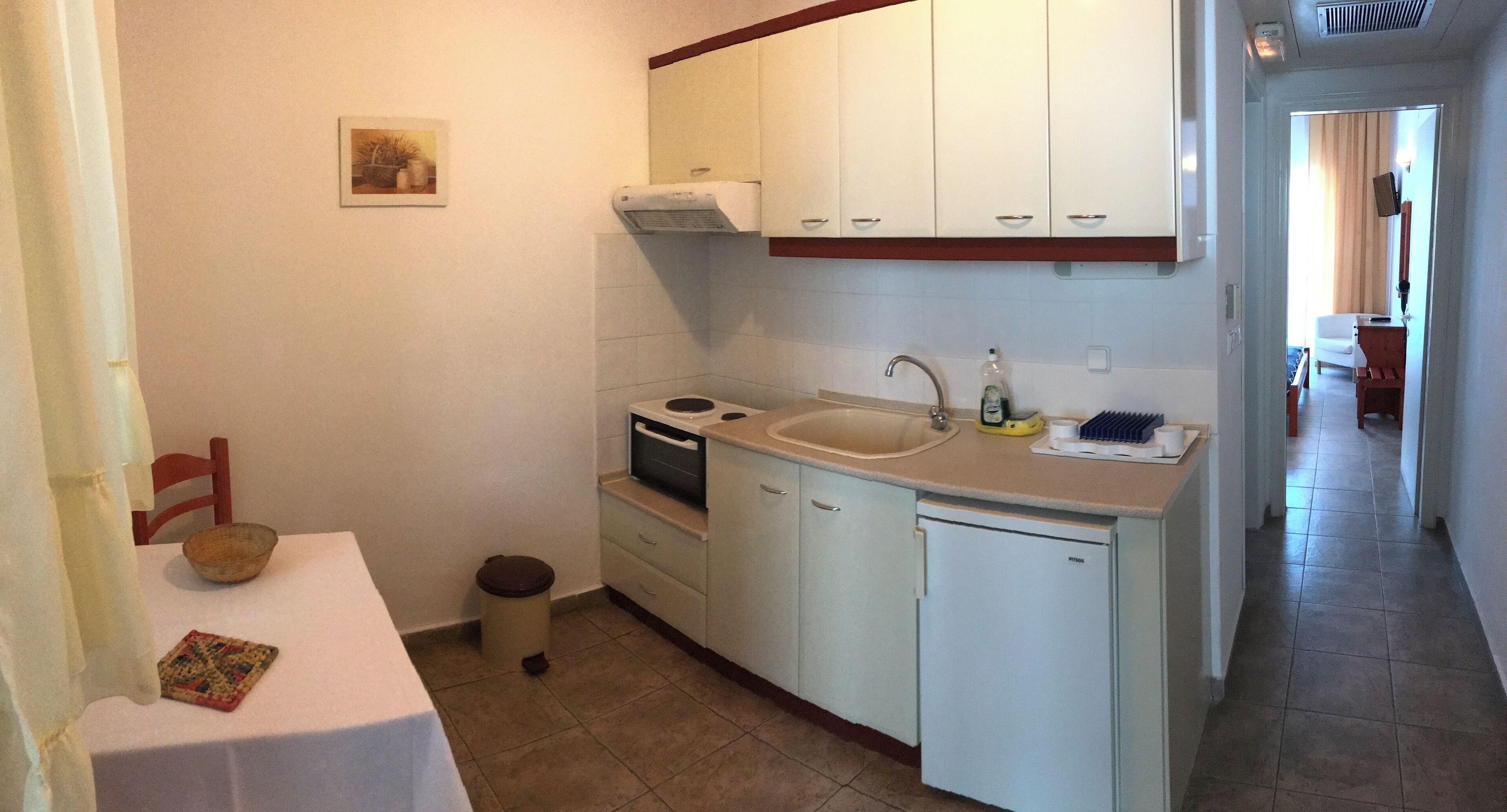 A compact kitchen with white cabinets, a sink, a stove, and a dishwasher. There is a small trash bin and a chair near the table.