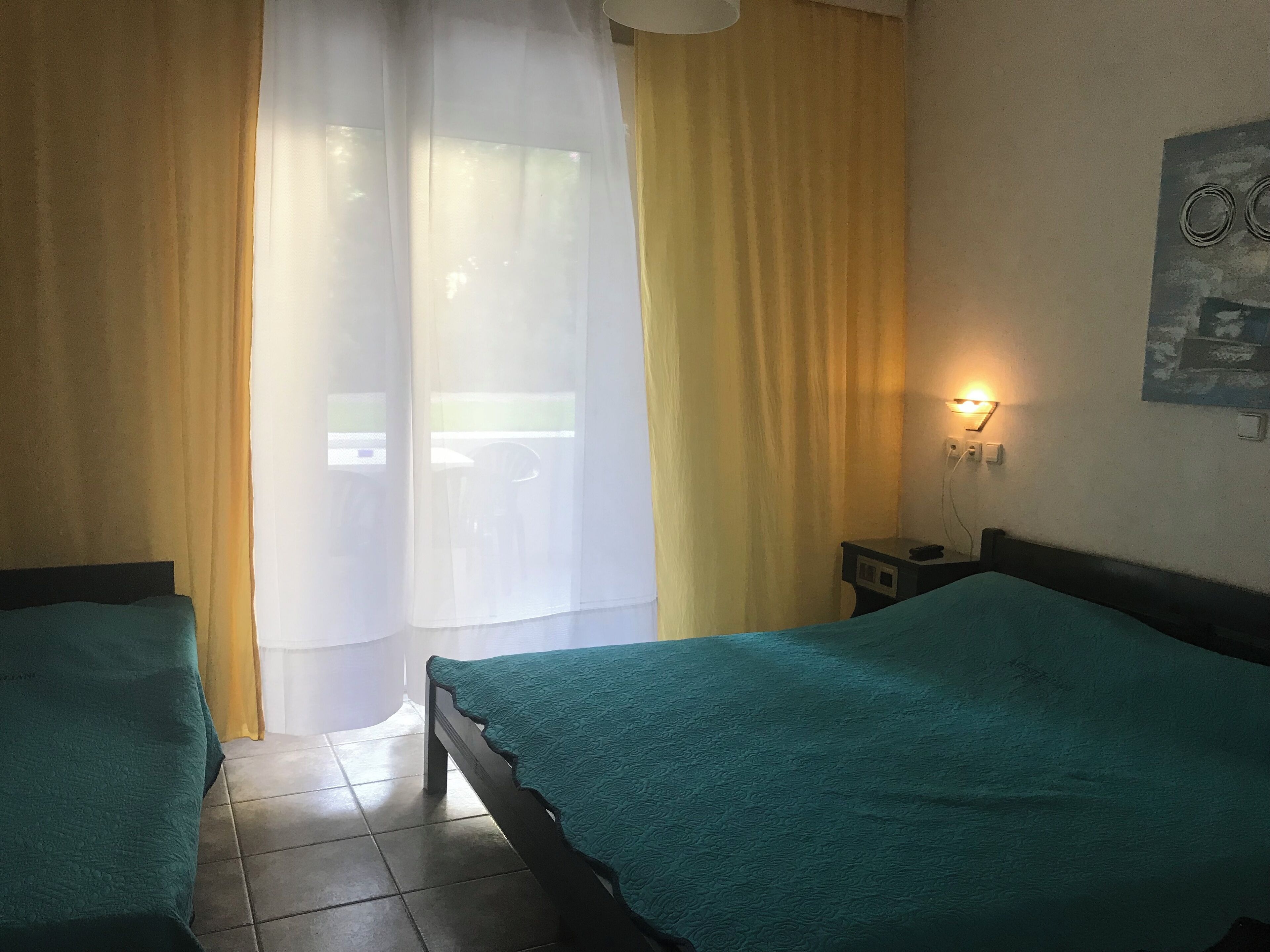 A room with two beds, a window with sheer curtains, a bedside table with a lamp, and a wall-mounted light switch.