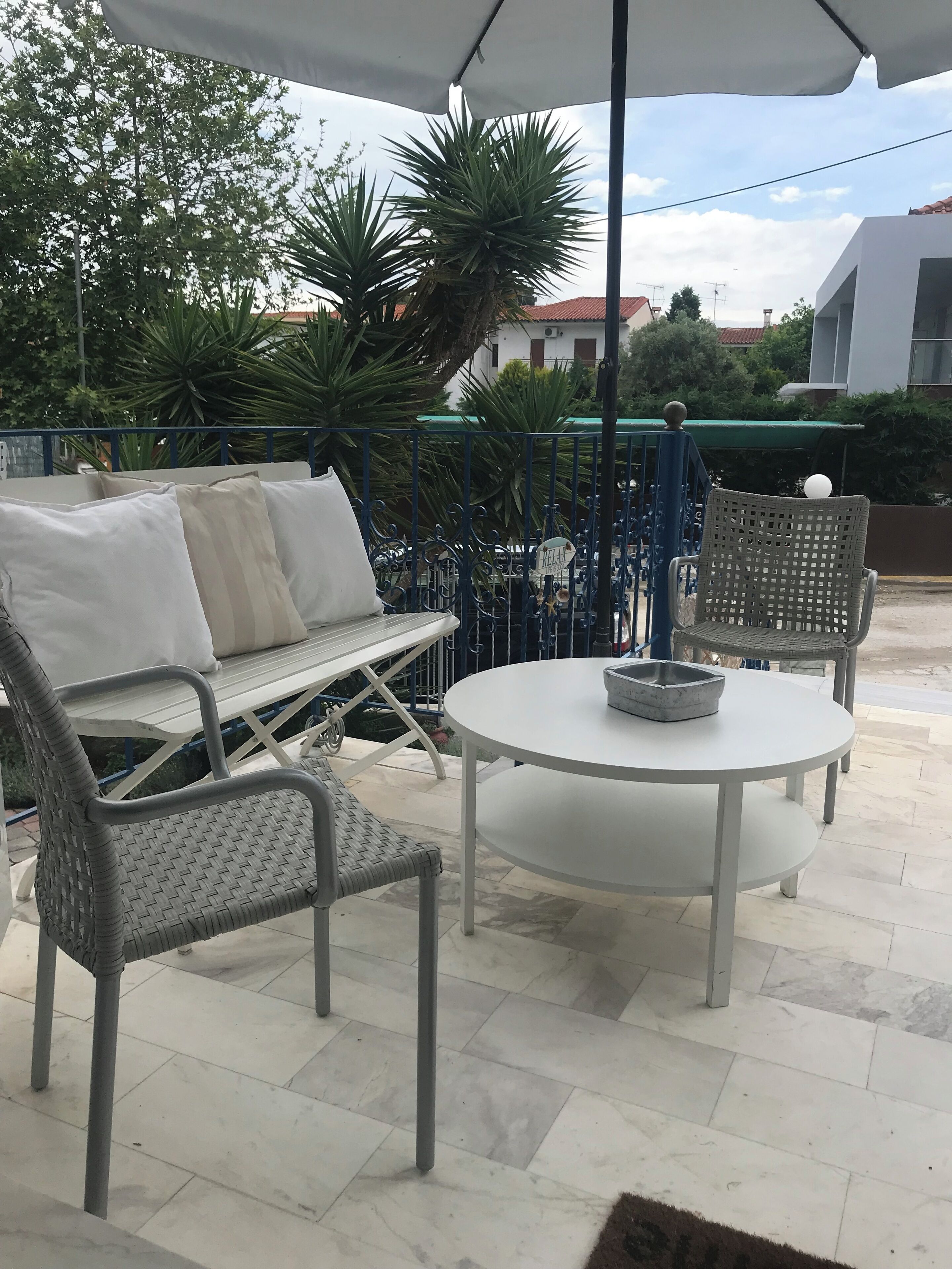 An outdoor patio area with a round table and two wicker chairs, a sofa with cushions, and a metal fence in the background.