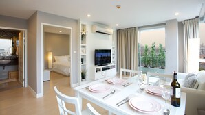A modern living room with a dining area, white furniture, a flat-screen TV, and a large window with a city view. - Cinnamon Hotel Bangkok (Bangkok)