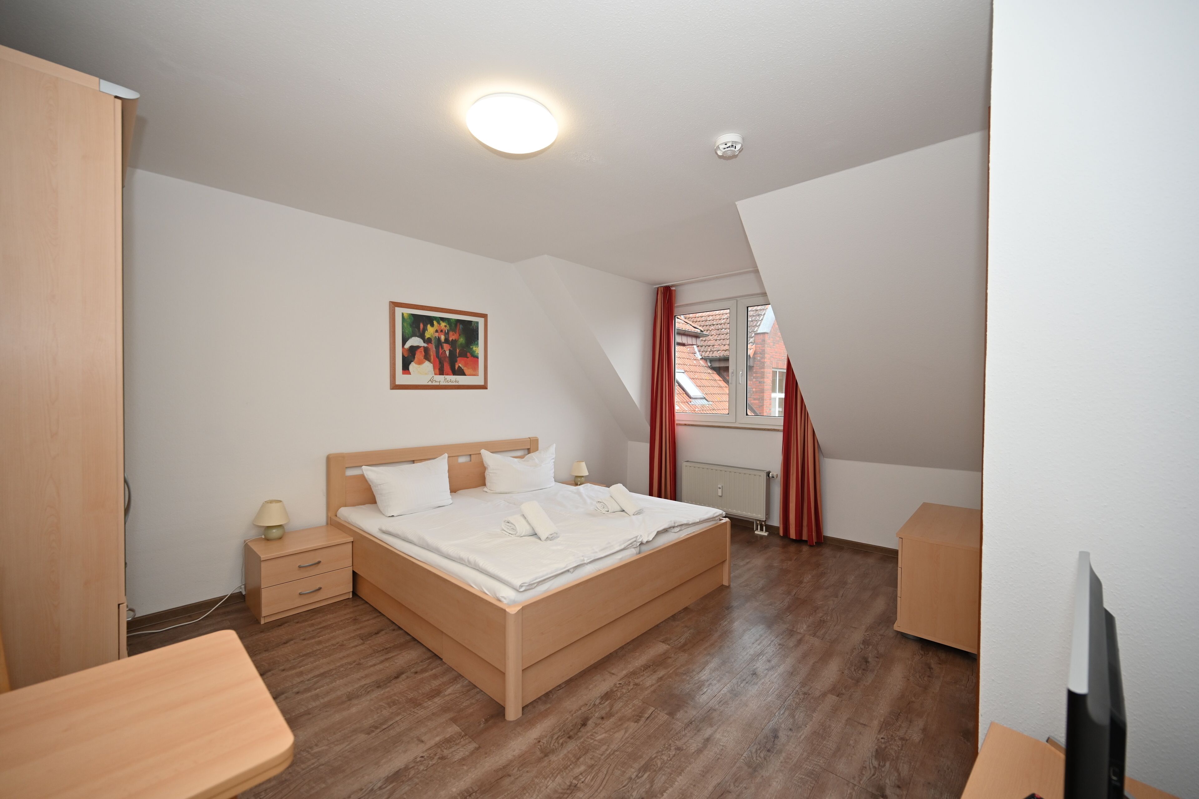 Ein Schlafzimmer mit einem hölzernen Bett, einem Nachttisch, einem Schreibtisch und einem Fenster mit roten Vorhängen.