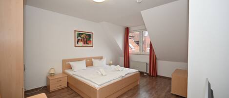 Ein Schlafzimmer mit einem hölzernen Bett, einem Nachttisch, einem Schreibtisch und einem Fenster mit roten Vorhängen.
