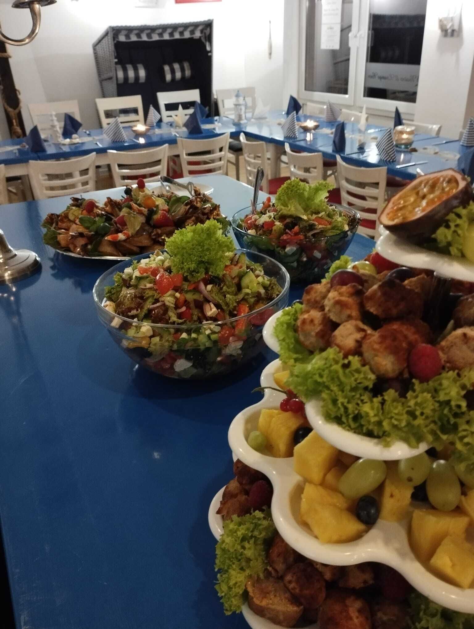 A buffet table with various dishes, including salads, fruits, and appetizers, set up in a dining area with white chairs and a blue tablecloth.