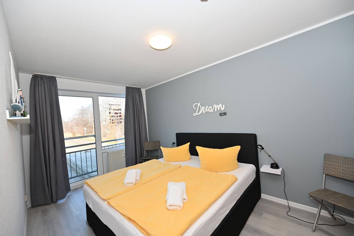 Ein modernes Schlafzimmer mit einem großen Bett, zwei Kissen, einem Nachttisch, einer Lampe und Blick ins Freie.
