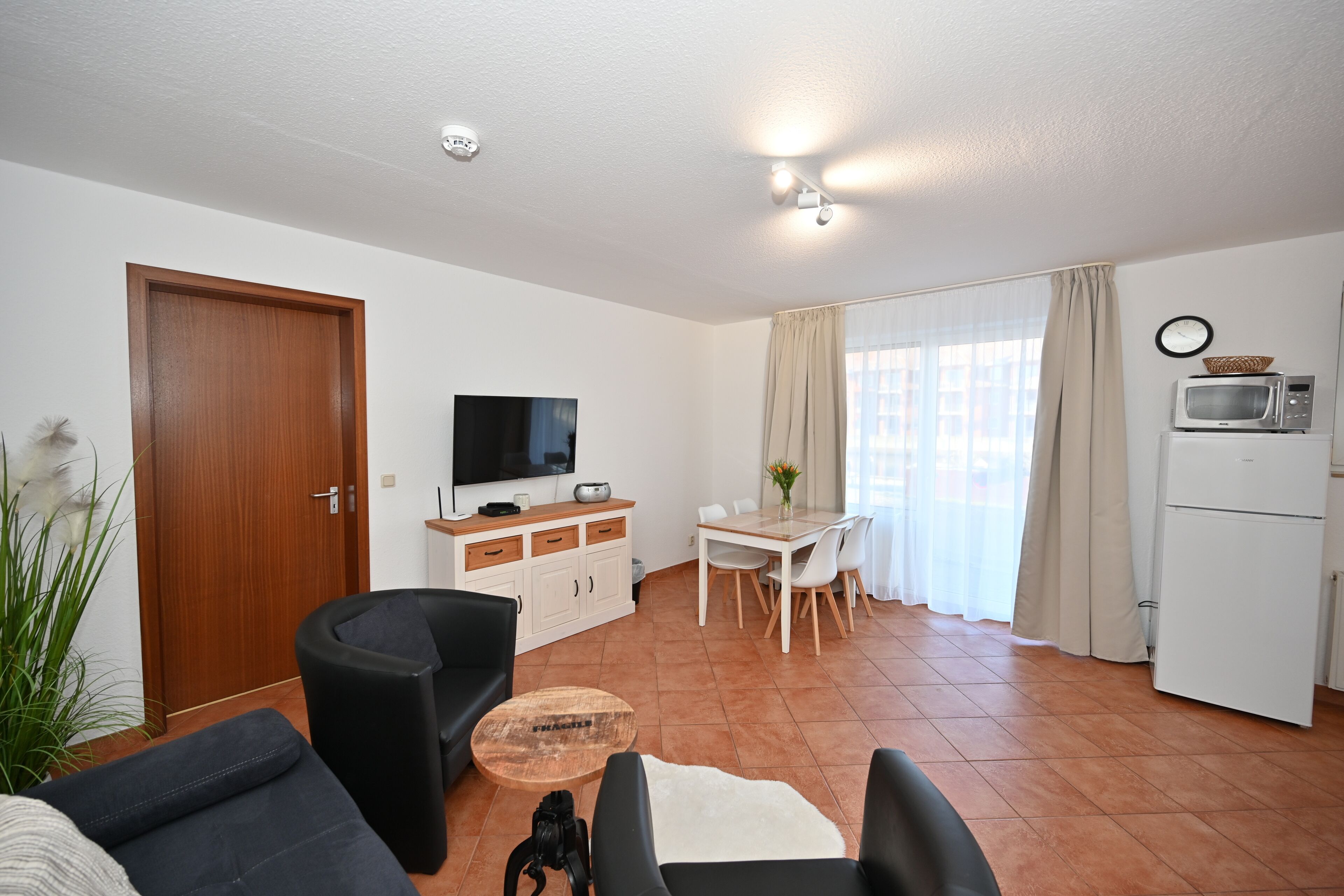 Una sala de estar con sofá, mesa de centro, televisor, mesa de comedor con sillas, una zona de cocina con microondas y una puerta.