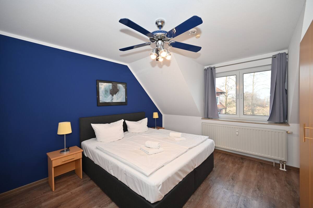 Ein Schlafzimmer mit Bett, Nachttisch, Lampe, Deckenventilator, Fenster mit Vorhängen und Heizkörper.