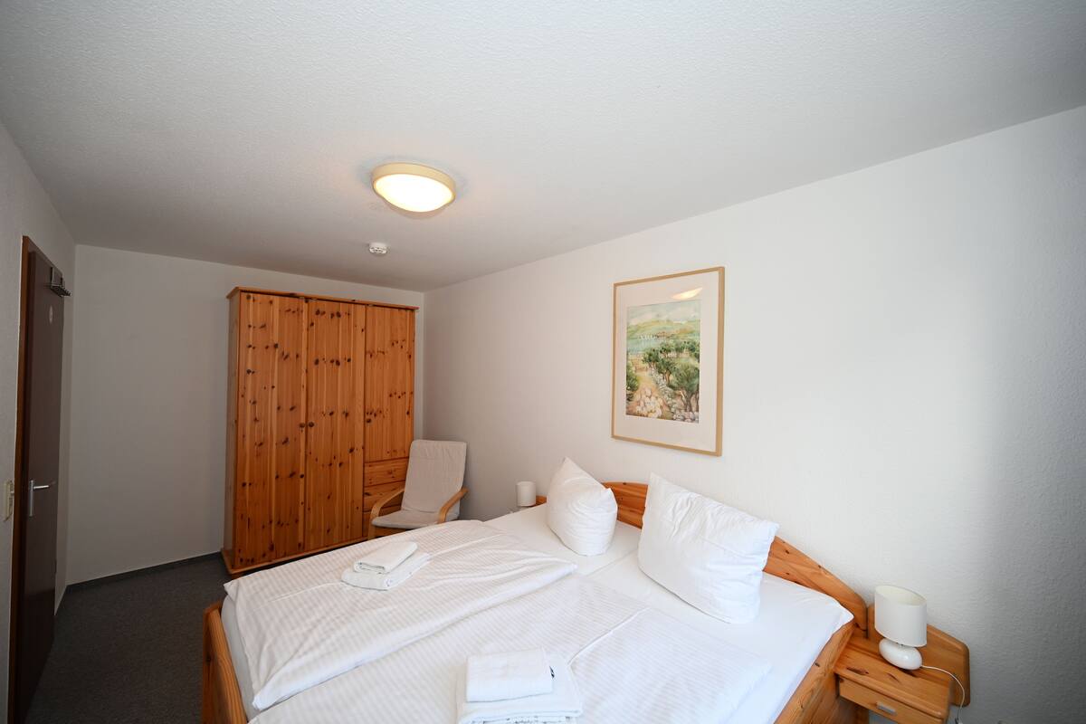 Ein Hotelzimmer mit einem Bett, einem Holzschrank, einem Sessel, einem Nachttisch mit Lampe und einem gerahmten Bild an der Wand.