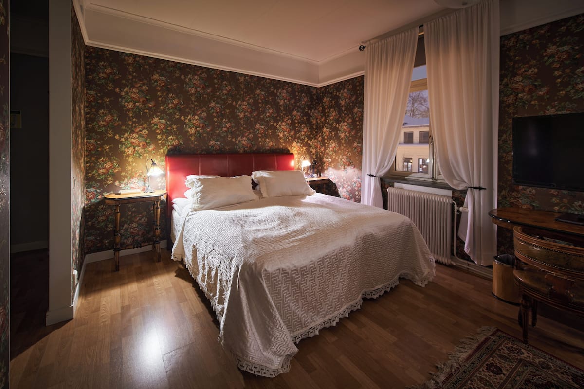 A bedroom with a large bed, a nightstand, a lamp, a dresser, a radiator, and a TV.