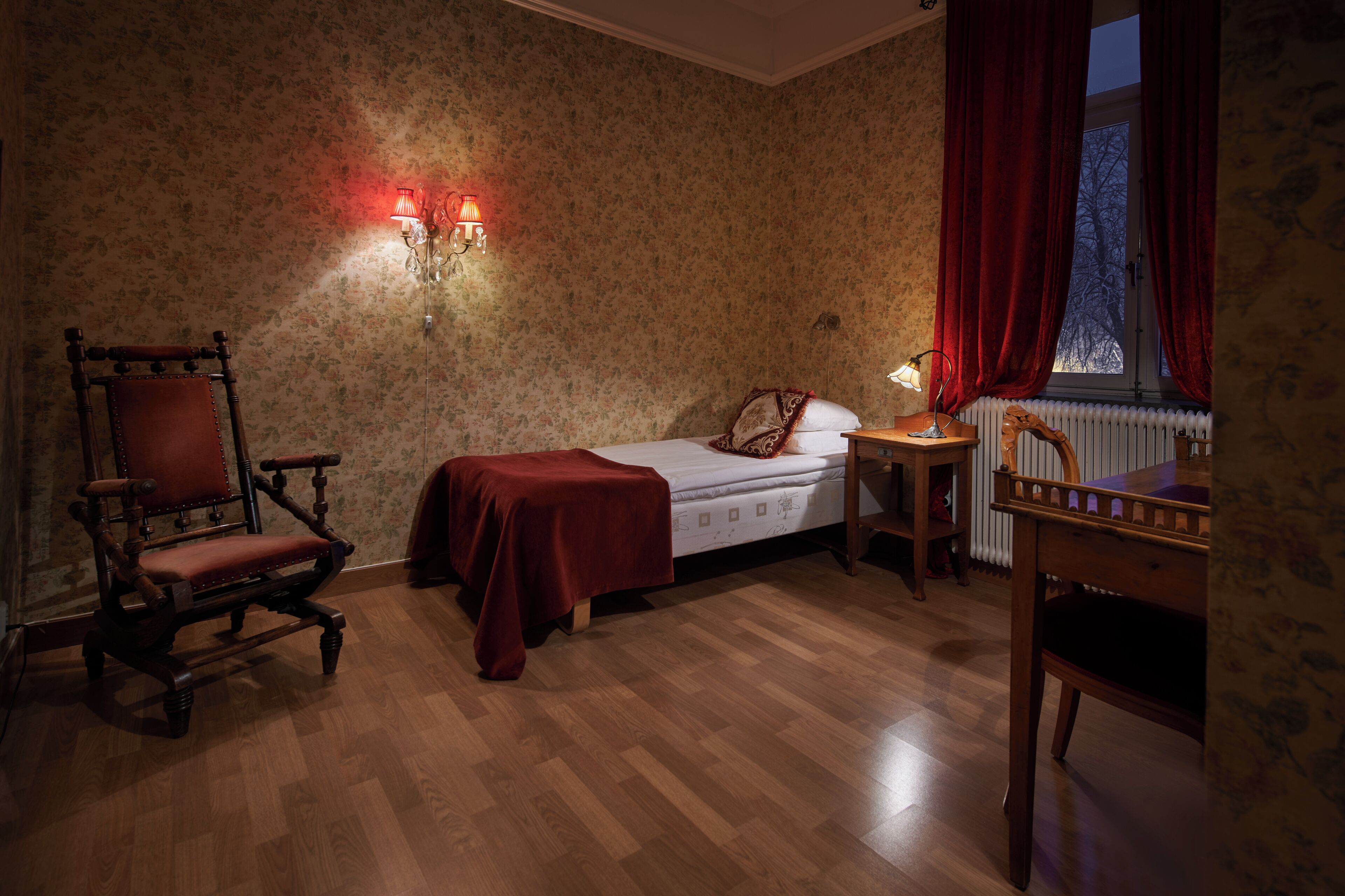 Une pièce avec un plancher en bois, un lit recouvert d’une couverture rouge, une chaise en bois, une table de chevet avec une lampe et une fenêtre ornée de rideaux rouges.