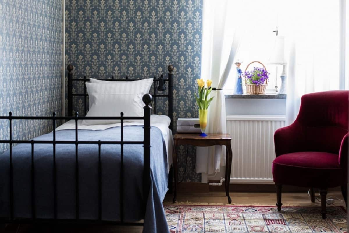 A bedroom with a bed, a red chair, a small table with a vase of flowers, and a window with curtains.
