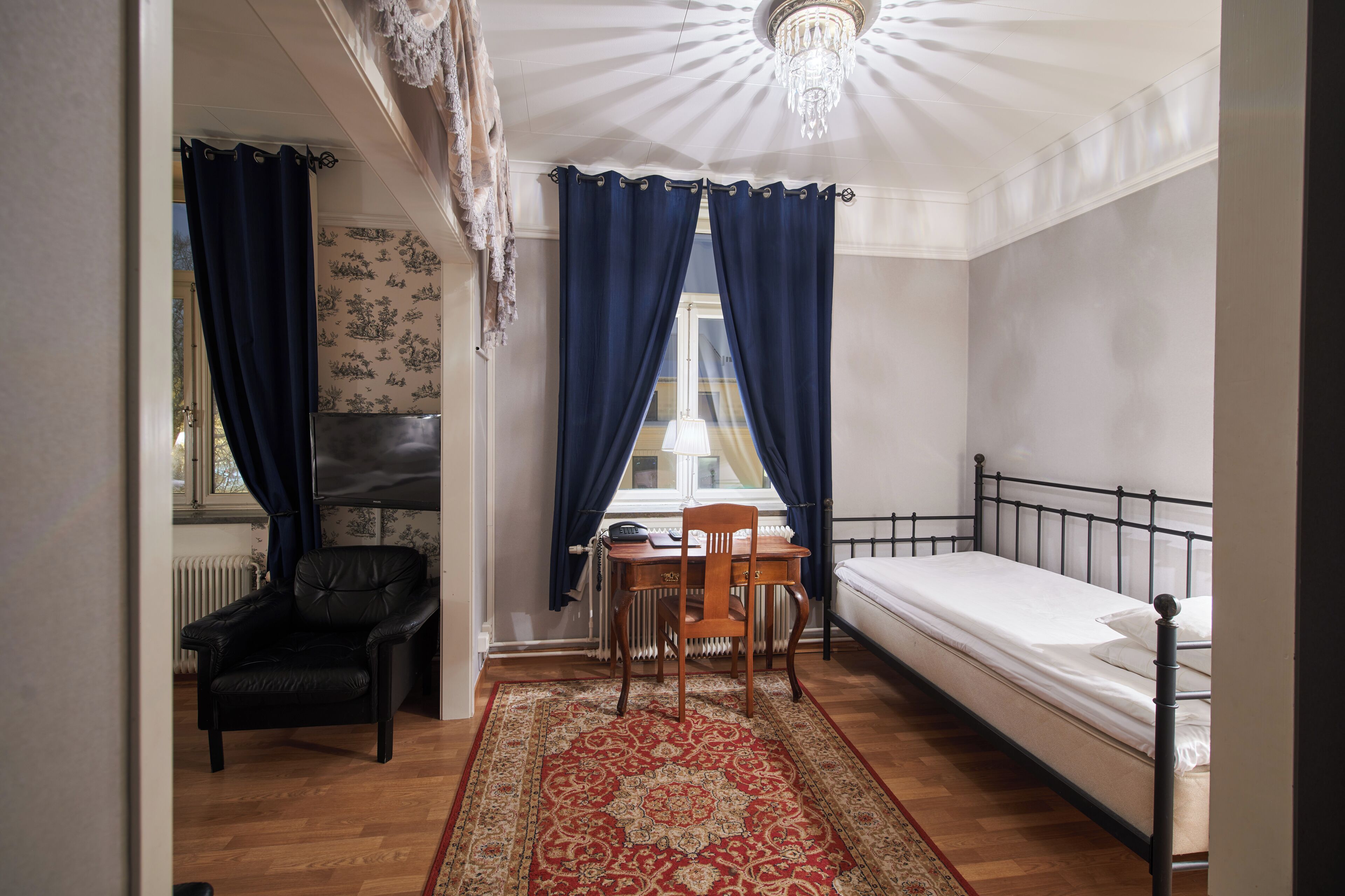 A small, well-lit room with a single bed, a wooden chair, a table, and a radiator.