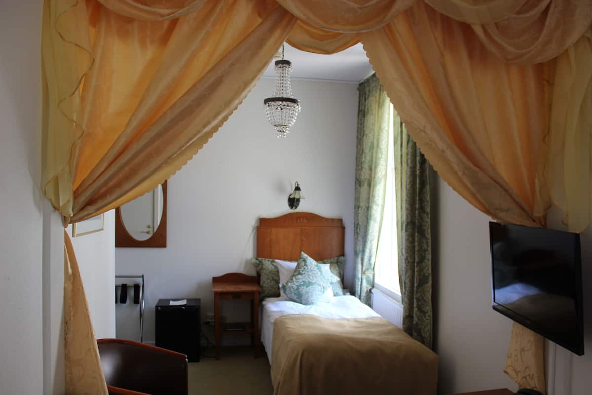 A hotel room with a bed, a bedside table, a TV mounted on the wall, and a window with curtains.