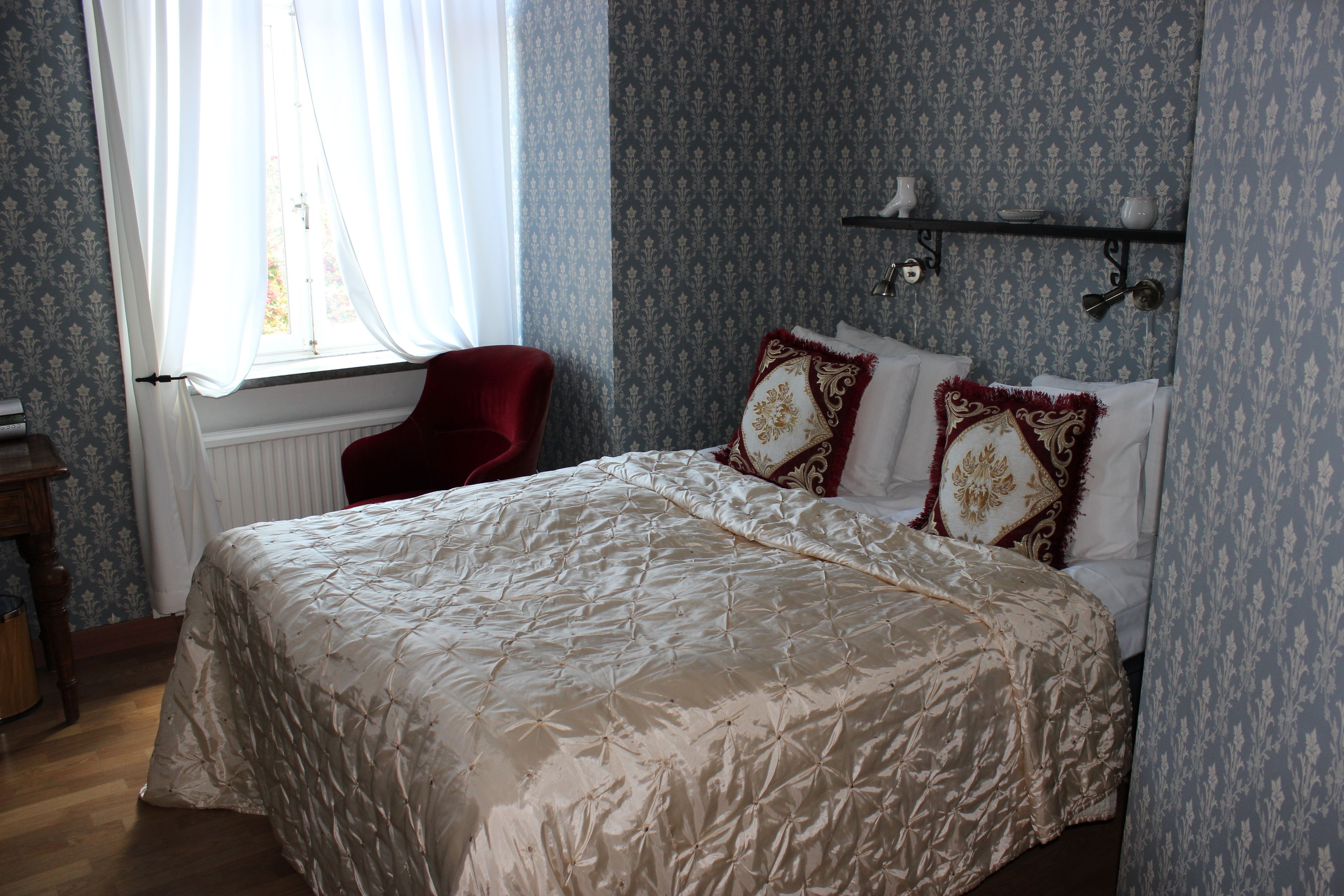 A neatly made bed with a patterned bedspread, a red chair, and a window with sheer curtains.