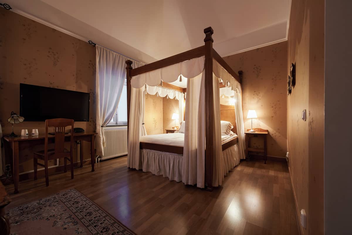 A four-poster canopy bed with a wooden frame and white bedding, a desk with a chair, a television, and a bedside table with a lamp.