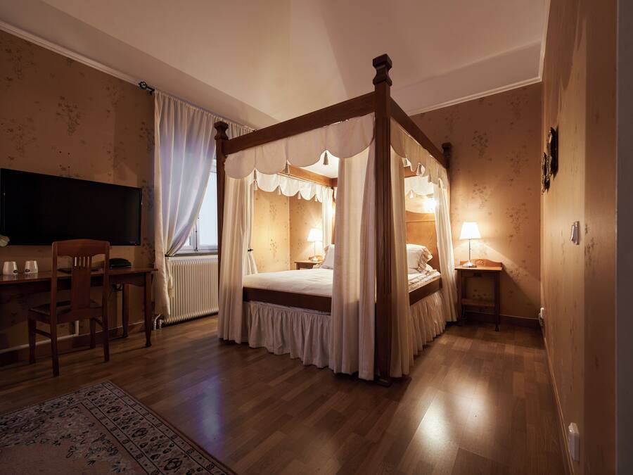 A four-poster canopy bed with a wooden frame and white bedding, a desk with a chair, a television, and a bedside table with a lamp.