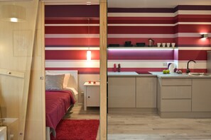 A modern bedroom with a red carpet and a wooden door. Adjacent is a kitchen with striped walls and a wooden countertop.