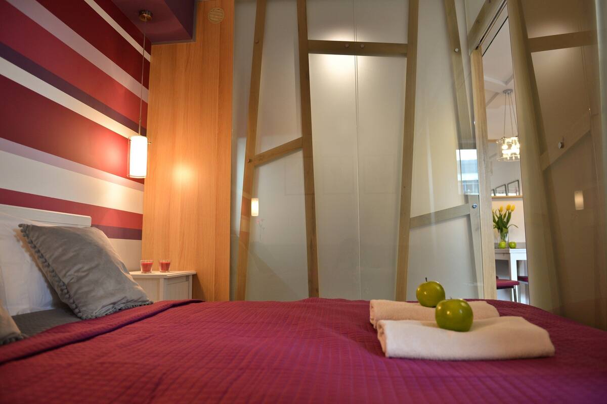 A hotel room with a red bedspread, a wooden headboard, a nightstand with a lamp, and a view of a dining area.