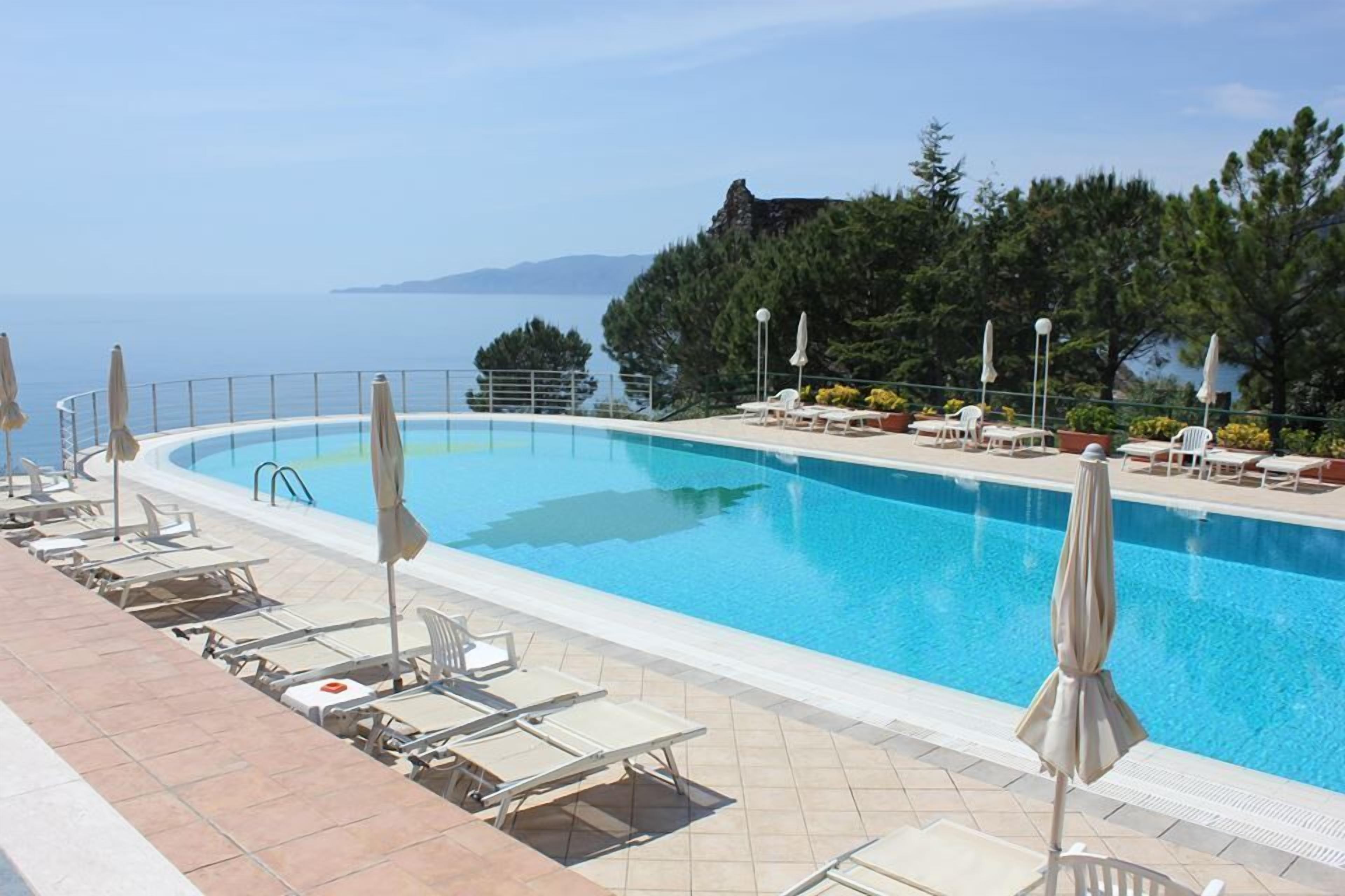 A pool area with lounge chairs and umbrellas, surrounded by trees and a view of the sea.