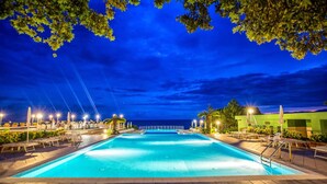 Ein Swimmingpool mit Liegestühlen und Sonnenschirmen, umgeben von Grünflächen und Blick auf das Meer in der Nacht.