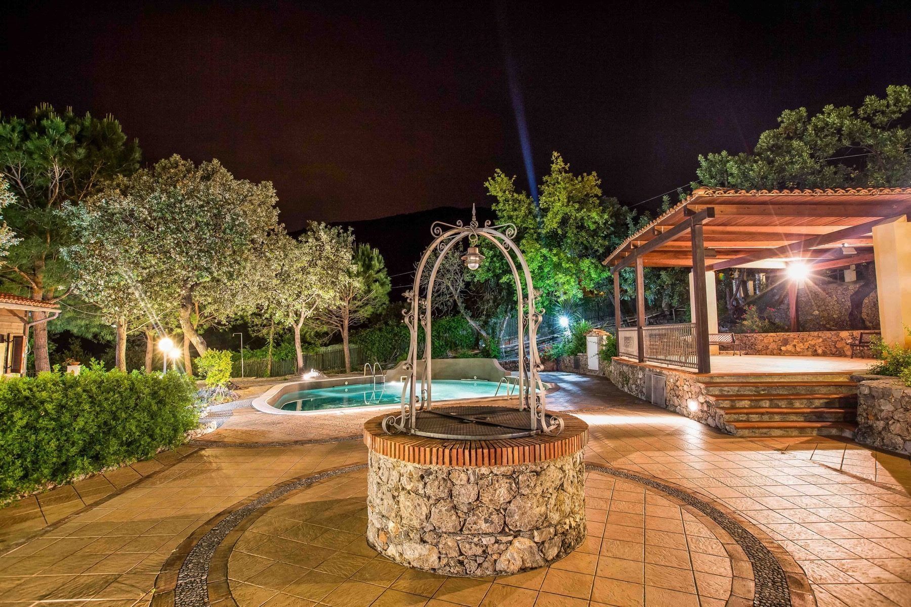 Un espace extérieur agrémenté d’une piscine bien éclairée, d’une fontaine en pierre, d’arbres et d’un espace de repos abrité.