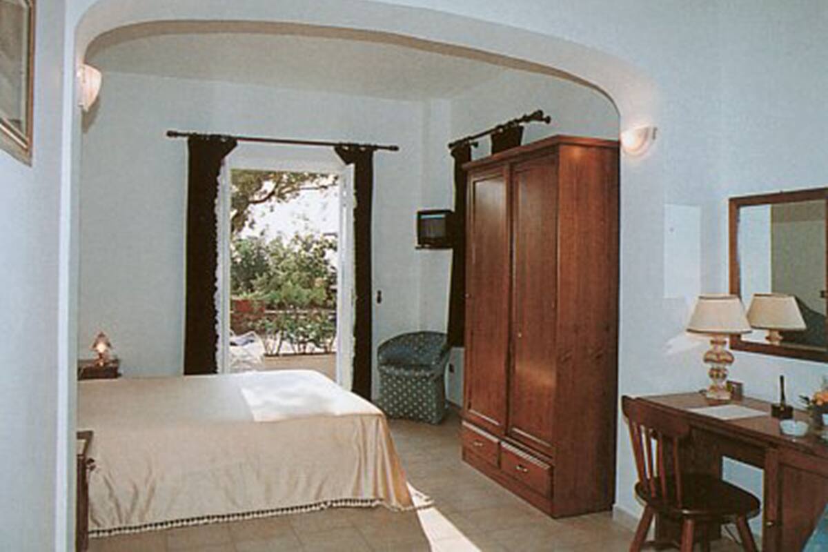 A bedroom with a bed, a wooden wardrobe, a desk with a lamp, and a window with curtains.