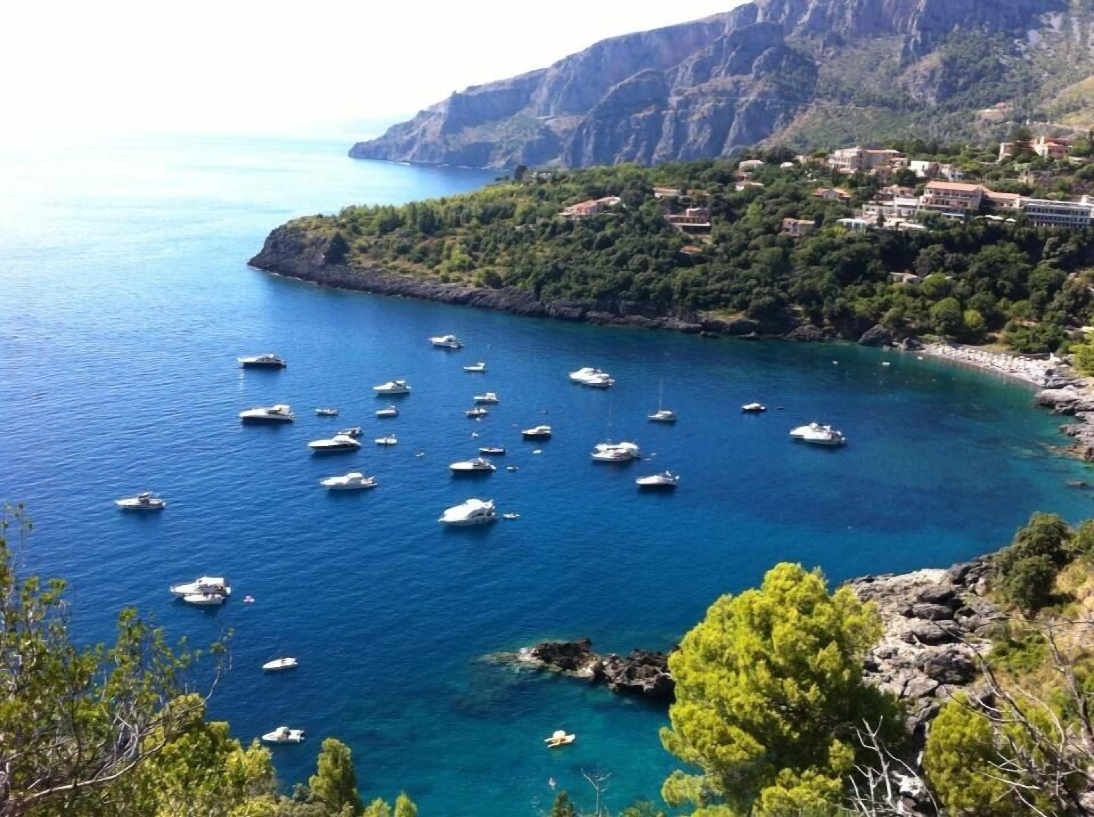 A coastal area with numerous boats anchored in clear blue waters, surrounded by rocky cliffs and dense greenery.