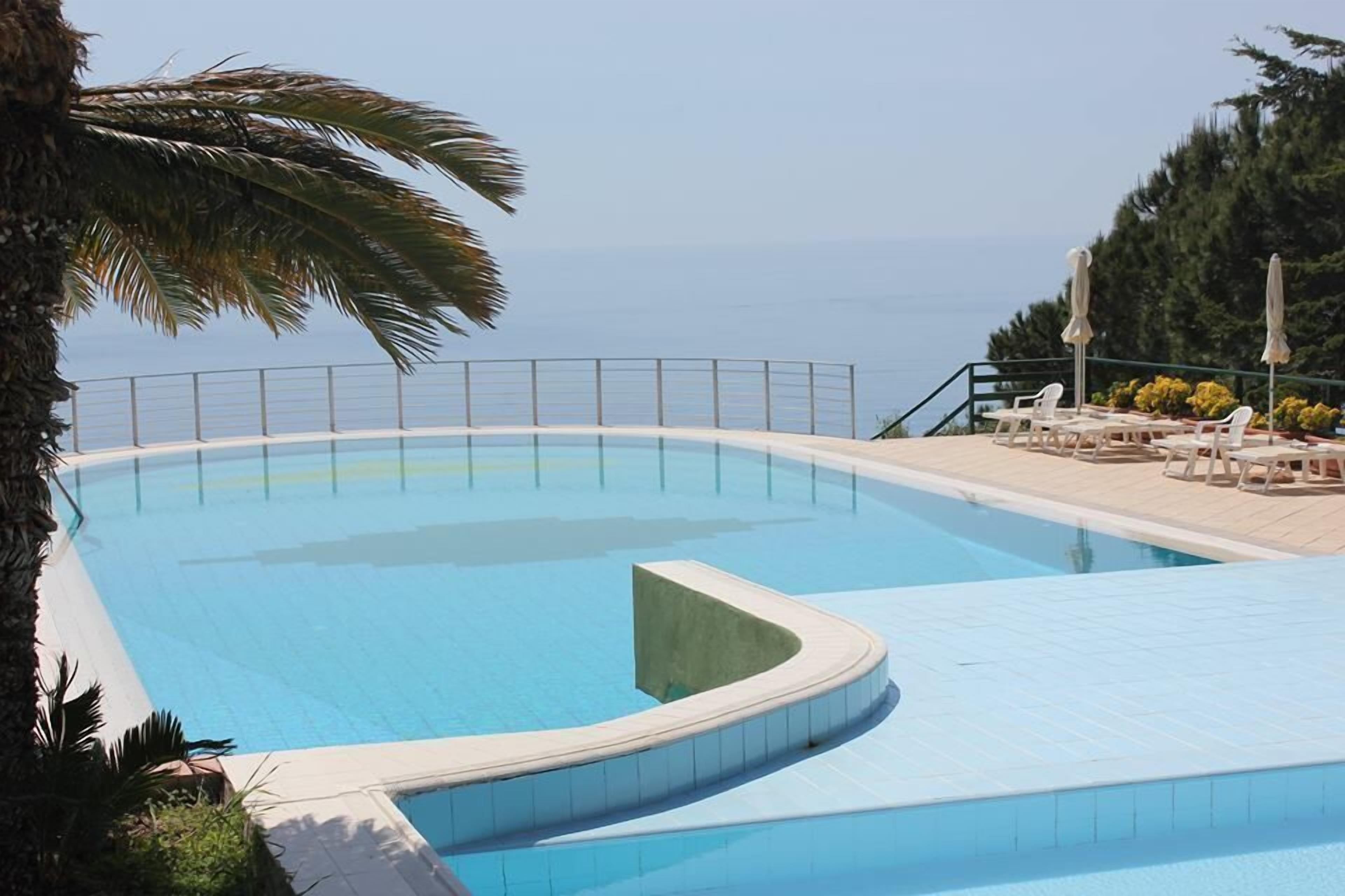 Ein Pool mit gewölbter Kante, umgeben von einer weißen Fliesenveranda und einem Metallgeländer. Auf der Veranda stehen Liegestühle und Sonnenschirme, und links ist eine Palme zu sehen.