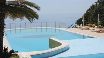 Ein Pool mit gewölbter Kante, umgeben von einer weißen Fliesenveranda und einem Metallgeländer. Auf der Veranda stehen Liegestühle und Sonnenschirme, und links ist eine Palme zu sehen.