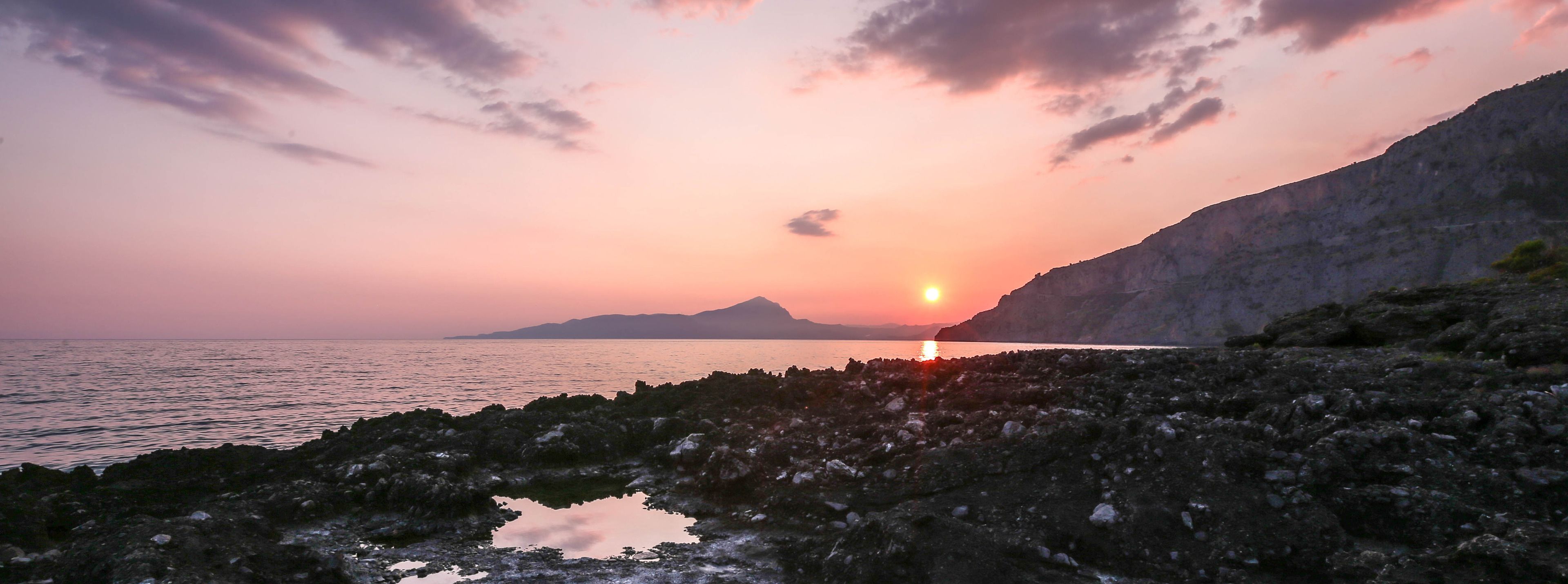 A sunset over a rocky coastline with a calm sea and distant mountains.