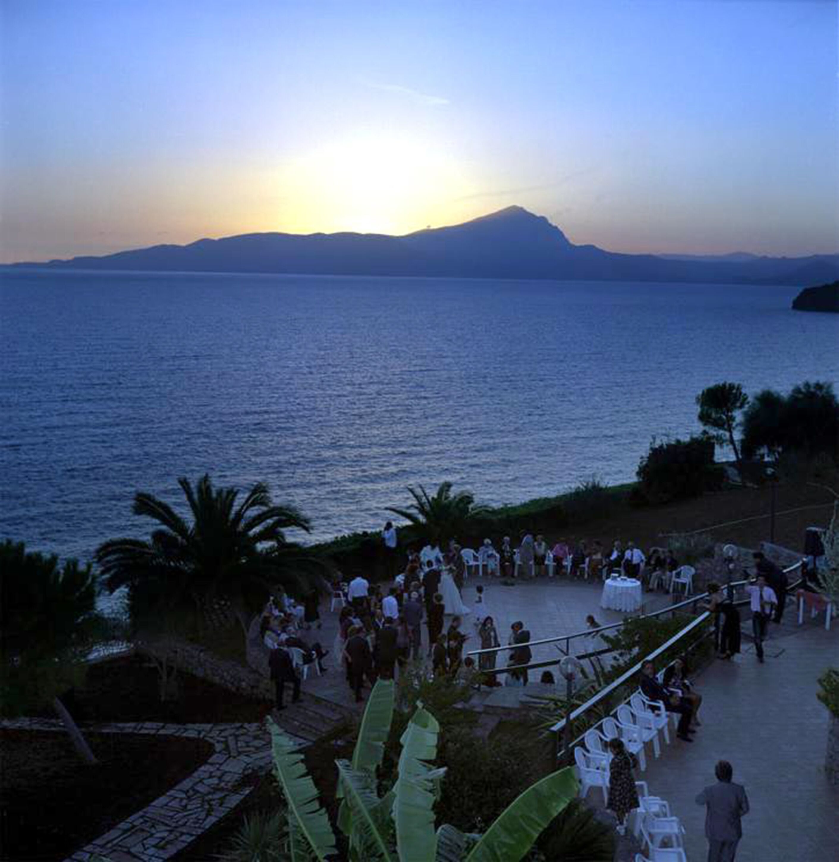 A coastal event with a crowd, a sunset, and a view of the sea and distant mountains.