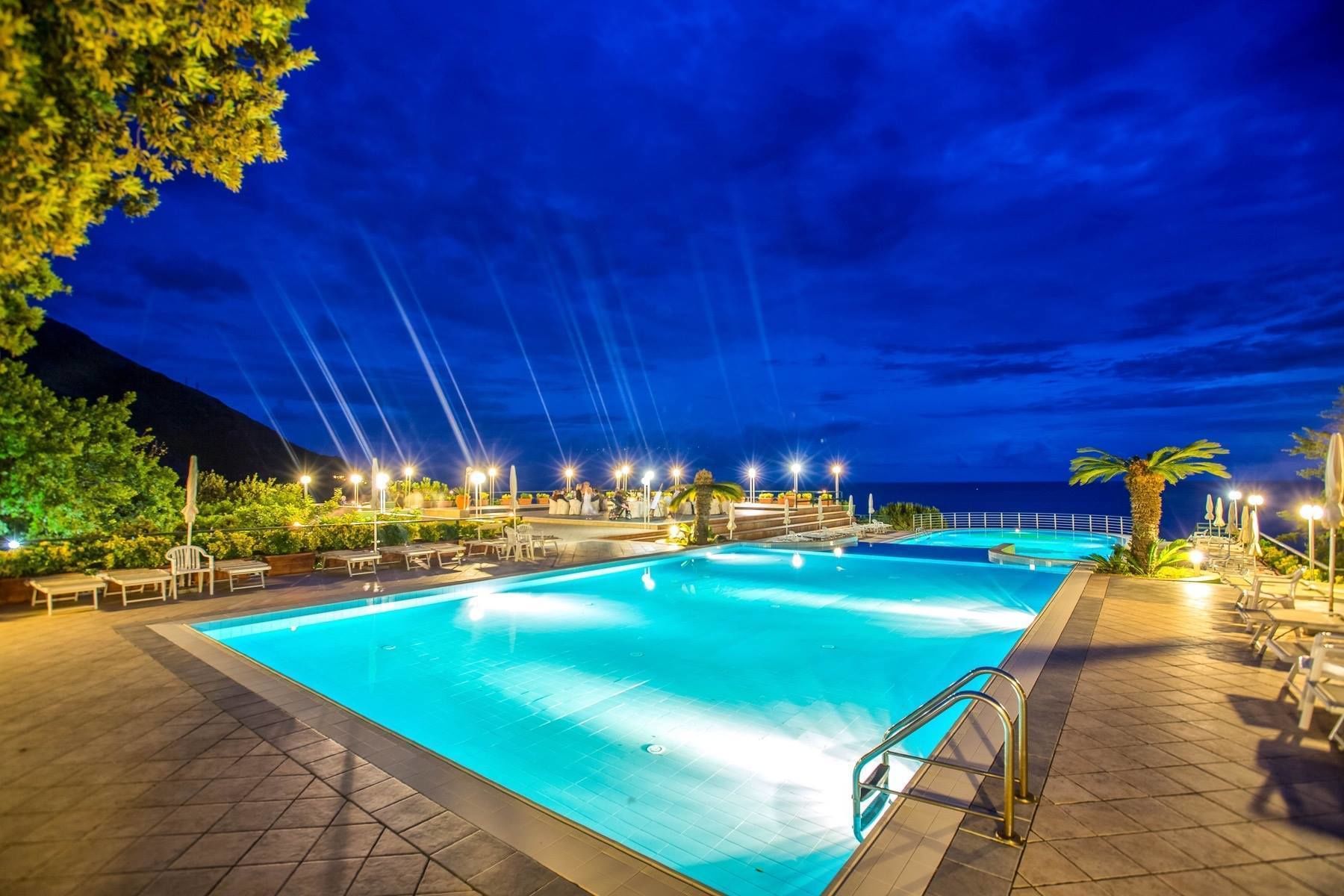 A nighttime swimming pool area with multiple pools, lounge chairs, and umbrellas, surrounded by trees and a view of the ocean.