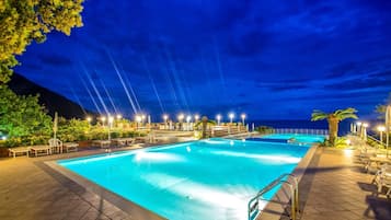 Zona de piscina nocturna con varias piscinas, sillas de descanso y sombrillas, rodeada de árboles y vistas al océano.