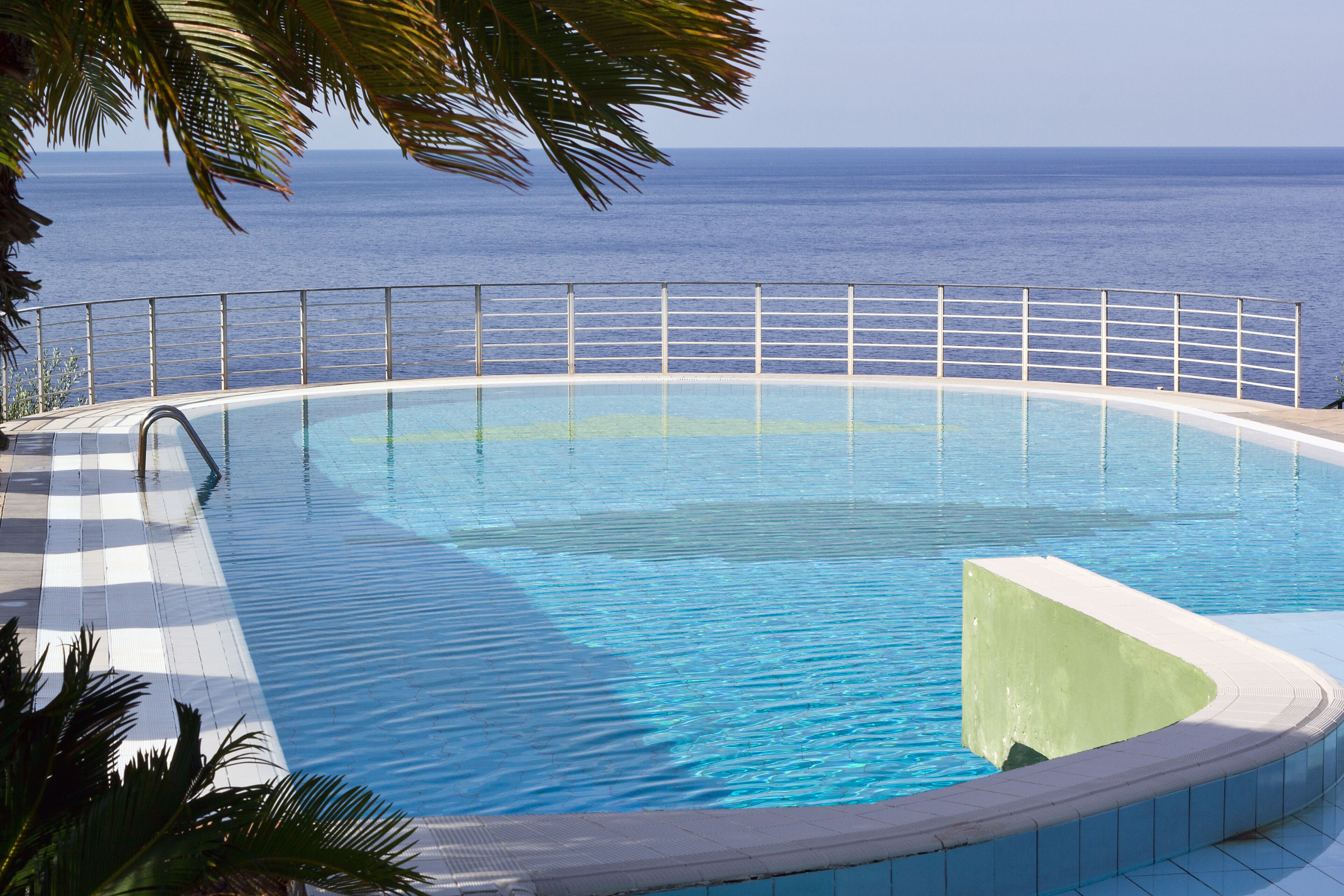 A pool with a view of the ocean, surrounded by a railing and palm trees.