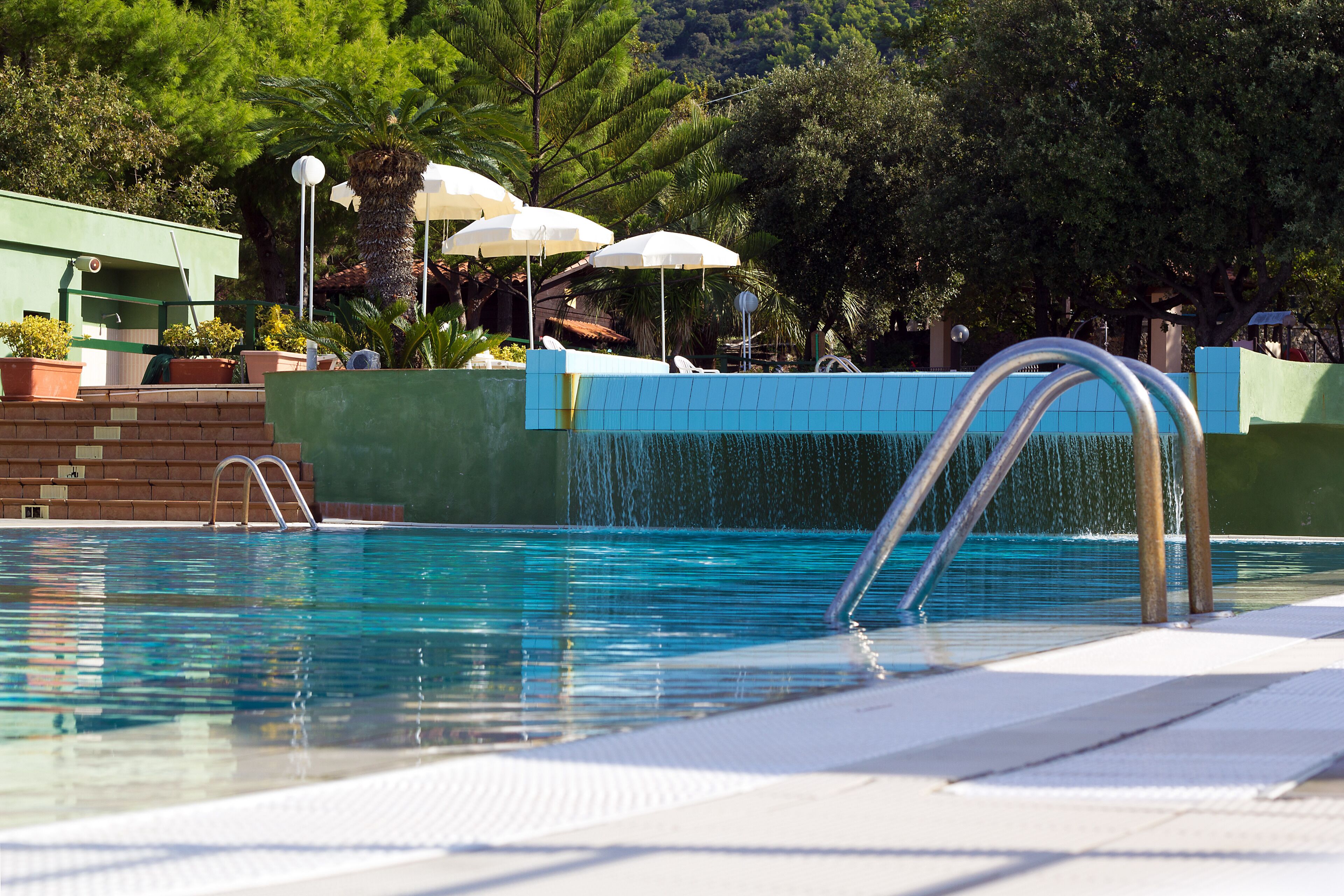 A swimming pool with a metal ladder, surrounded by potted plants and umbrellas.